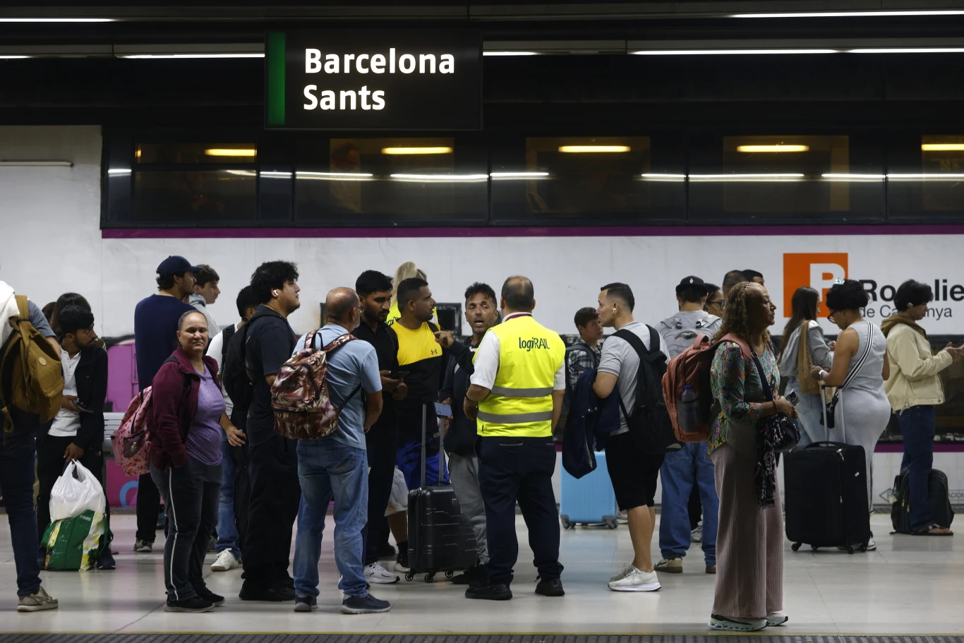 Grupa osób z bagażami czeka na peronie stacji metra Barcelona Sants, wśród nich pracownik w żółtej kamizelce rozmawia z podróżnymi.