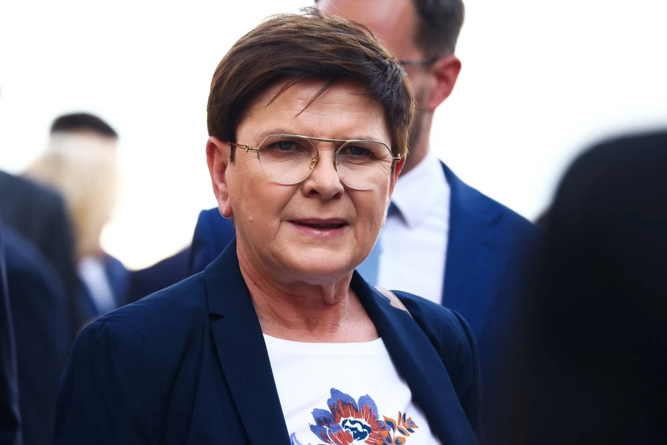 Beata Szydło, była premier Polski, obecnie europosłanka i wiceprezeska PiS-u Kobieta w krótkich włosach i okularach ubrana w granatową marynarkę z białą bluzką z kolorowym wzorem, stojąca na tle innych osób ubranych w garnitury.