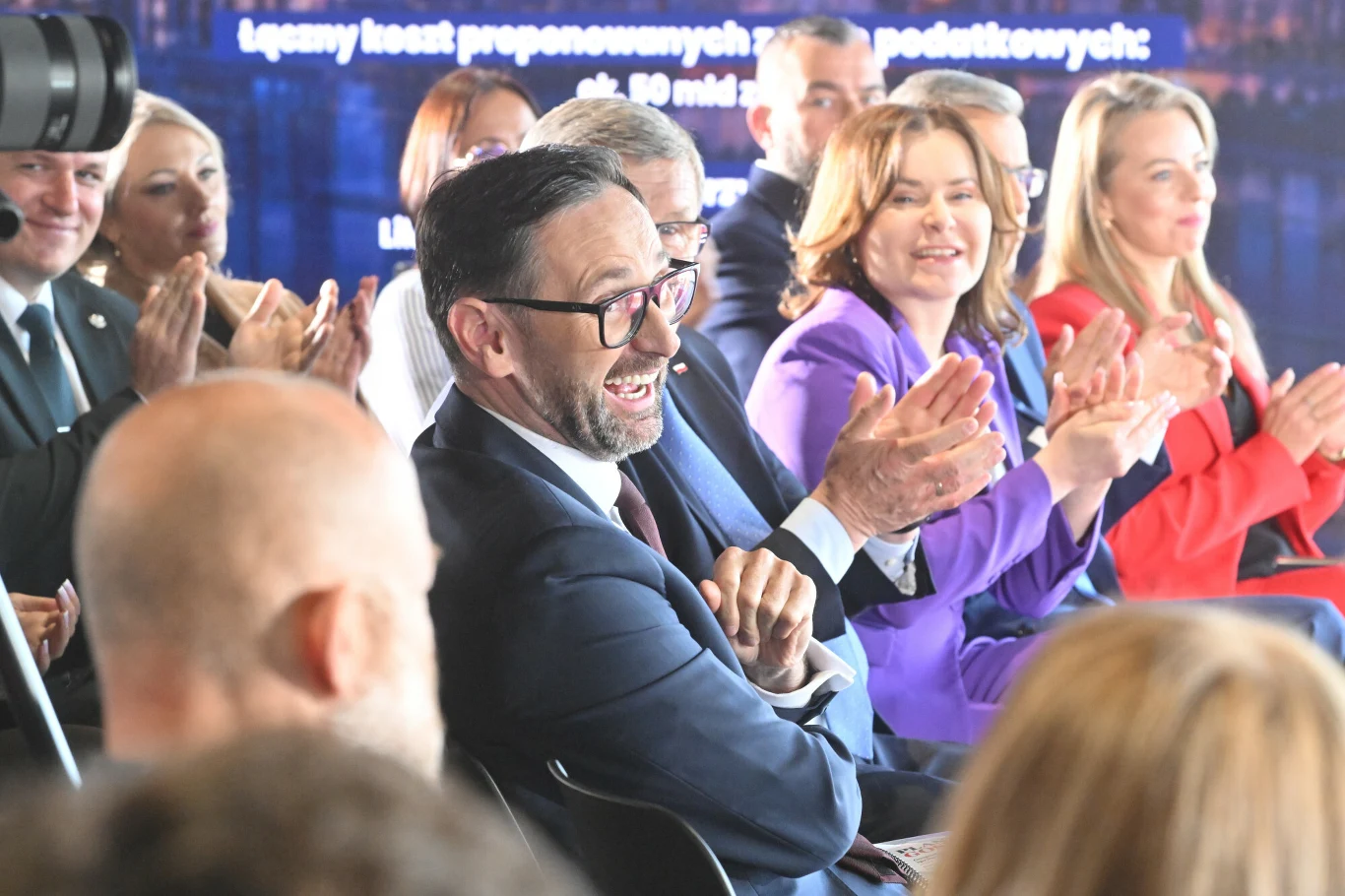 Daniel Obajtek, europoseł PiS-u i były prezes Orlenu Grupa elegancko ubranych osób siedzących w rzędzie, część z nich uśmiecha się i bije brawo, atmosfera spotkania oficjalnego lub prezentacji.
