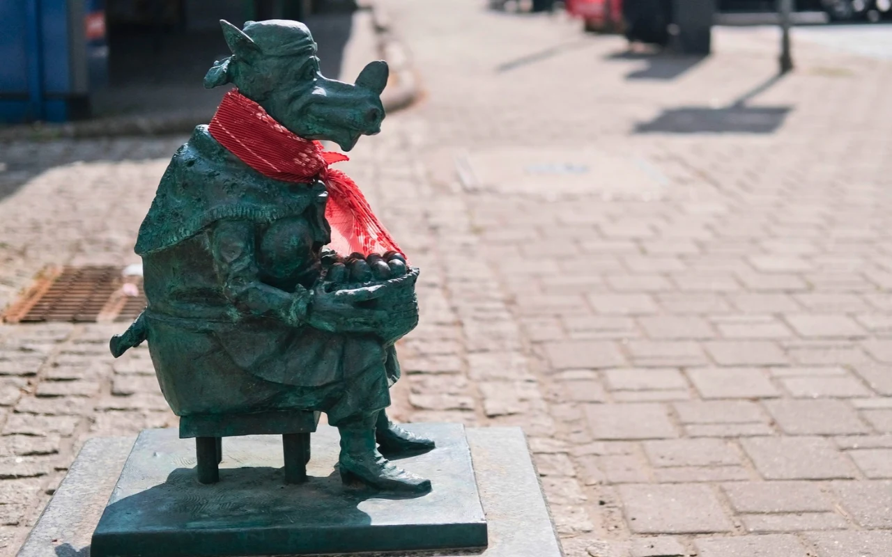 Smoczyca Przekupka w Krakowie Brązowa figurka smoka, z czerwonym szalikiem na szyi, trzymającego koszyk z jabłkami, ustawiona na chodniku.