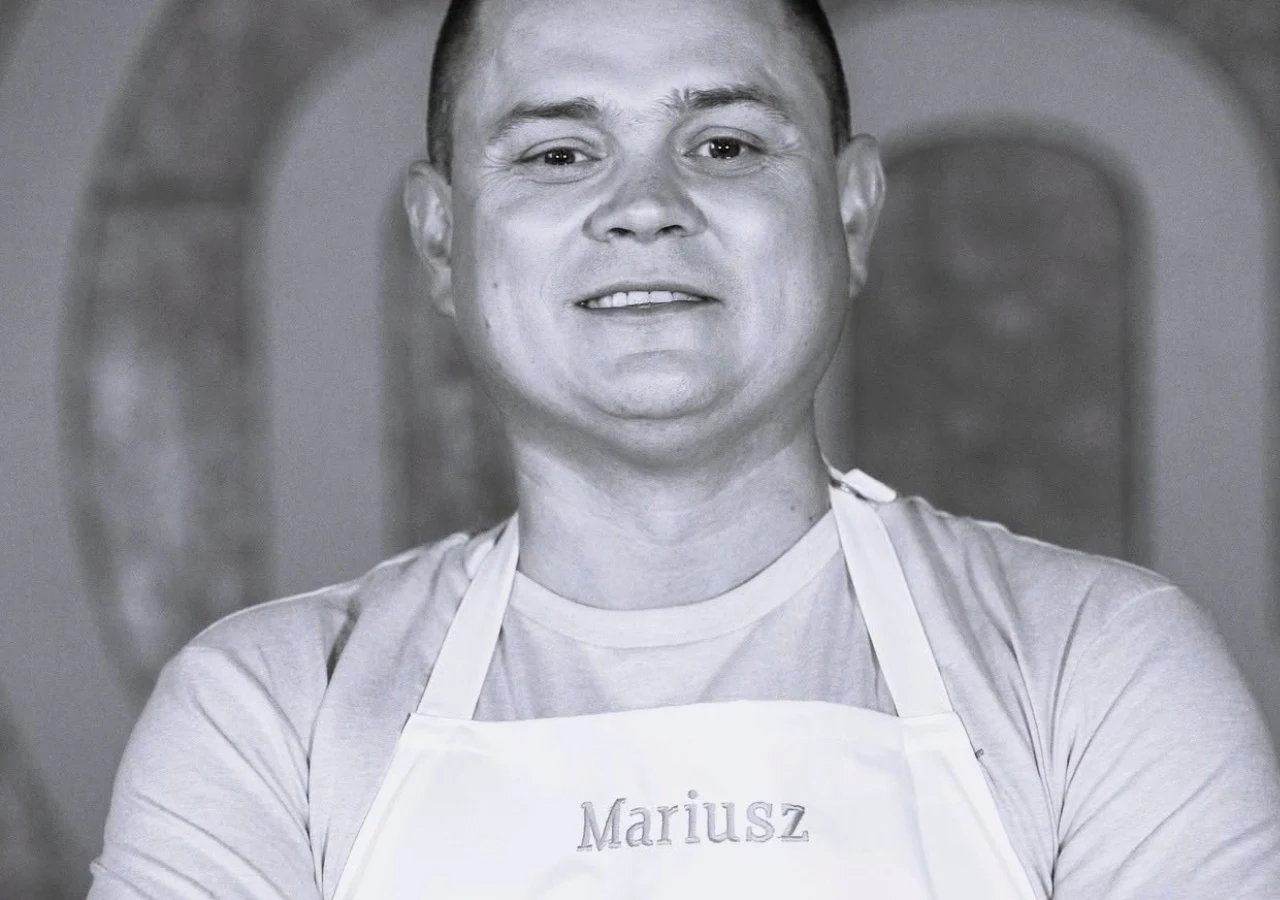 Mariusz Komenda w programie "MasterChef" Mężczyzna w jasnej koszulce i fartuchu z imieniem „Mariusz”, uśmiechnięty, sfotografowany od przodu w neutralnym otoczeniu.