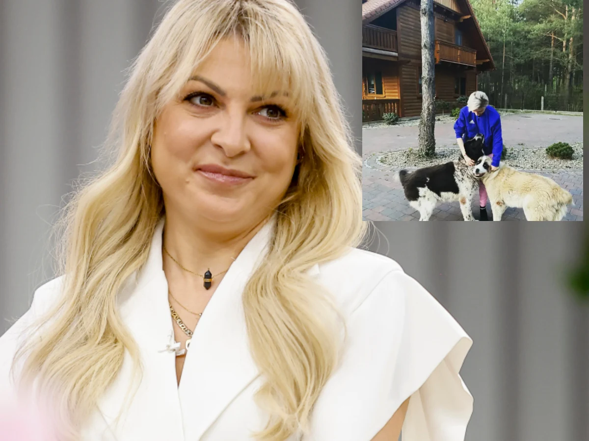 Blondynka w białej marynarce z delikatnym makijażem uśmiecha się lekko, a obok kobieta głaszcze dwa duże psy na tle drewnianego domu w lesie.