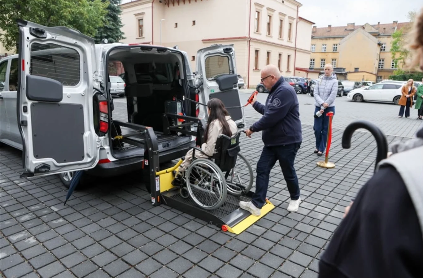 Taxi Help w Tarnowie. Z usługi mogą skorzystać m.in. seniorzy. Kobieta na wózku inwalidzkim korzysta z windy samochodu Taxi Help w Tarnowie, asystowana przez mężczyznę.