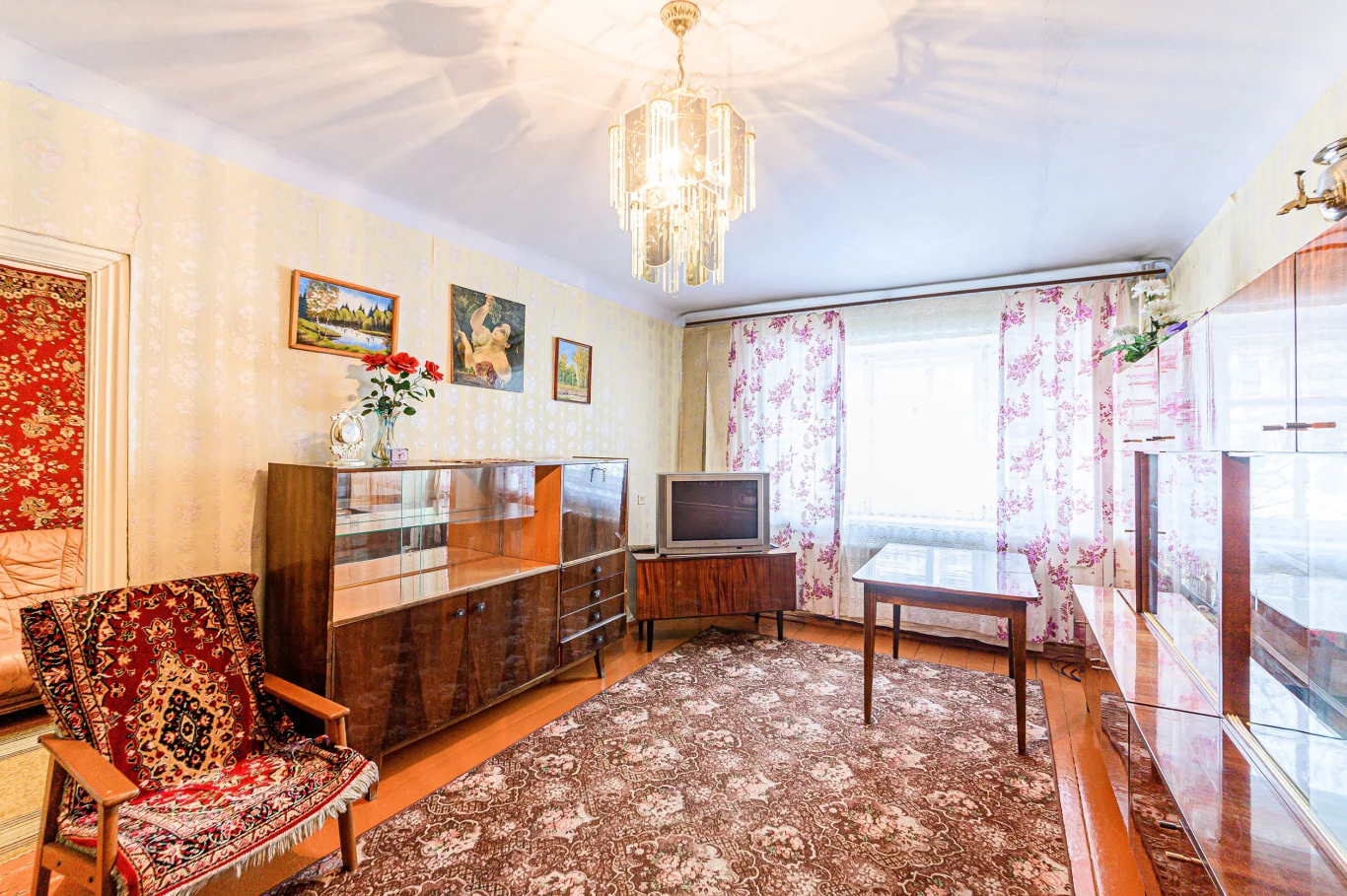 We wnętrzach z czasów PRL-u znajdziemy elementy, które możemy wykorzystać w stylu grandma chic. Pokój urządzony w stylu lat 80., z żyrandolem, segmentem, telewizorem na niskim stoliku, dywanem w kwiatowy wzór i fotelem.