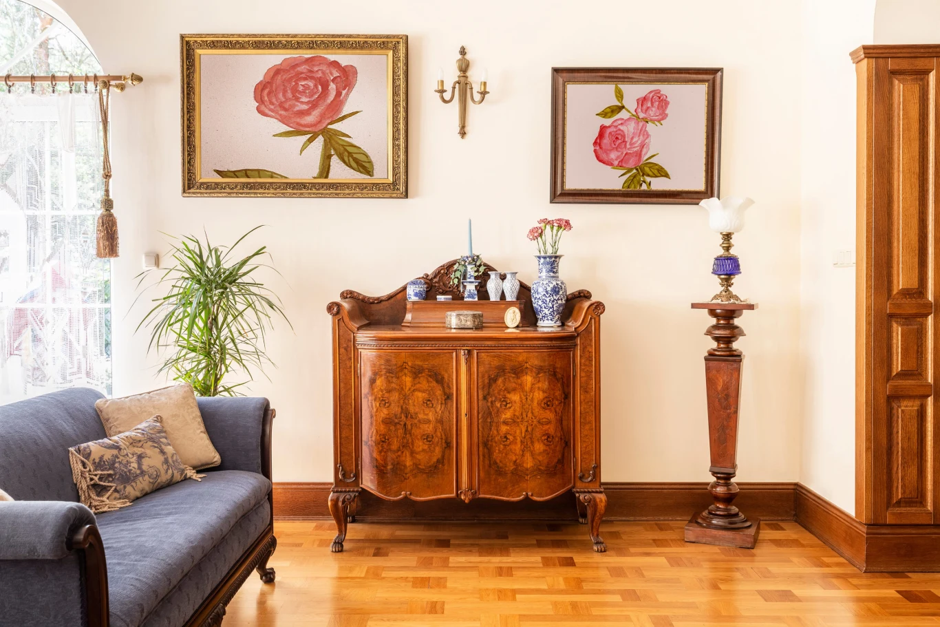 Do nowoczesnego wnętrza można wprowadzić akcenty w stylu retro. Stylowy salon z zabytkową komodą, na której stoją porcelanowe wazony i dekoracyjne przedmioty, otoczony klasycznymi obrazami z motywami kwiatowymi, drewnianą podłogą i elegancką sofą po lewej stronie.