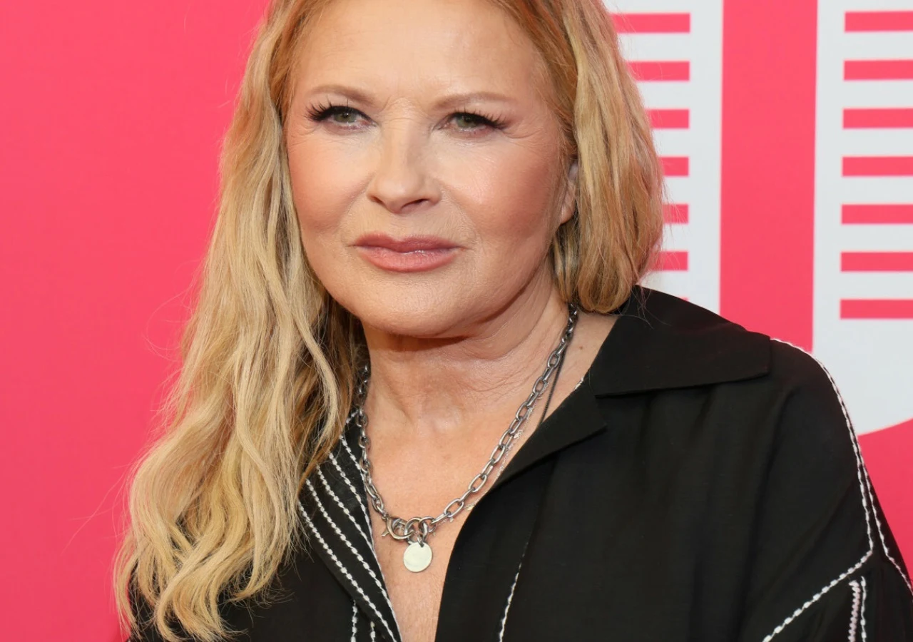 Urszula Piosenkarka Urszula z długimi blond włosami, w czarnej koszuli i z łańcuszkami na szyi, pozuje na różowym tle.