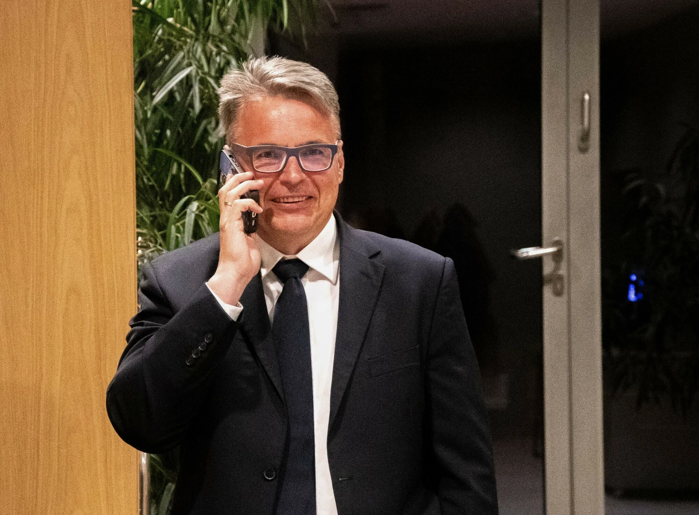 Mężczyzna w garniturze i okularach rozmawia przez telefon, stojąc w nowoczesnym wnętrzu z roślinami w tle.