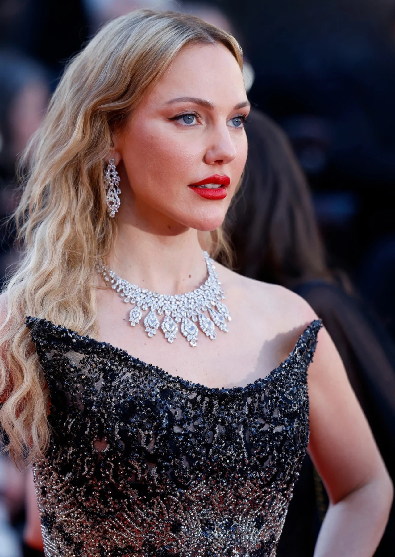 Meryem Uzerli zachwyca Kobieta w efektownej, czarno-srebrnej sukni bez ramiączek, z długimi blond falowanymi włosami, ozdobiona dużą, lśniącą biżuterią – kolczykami i naszyjnikiem, patrzy zdecydowanie w bok na tle rozmytego otoczenia.