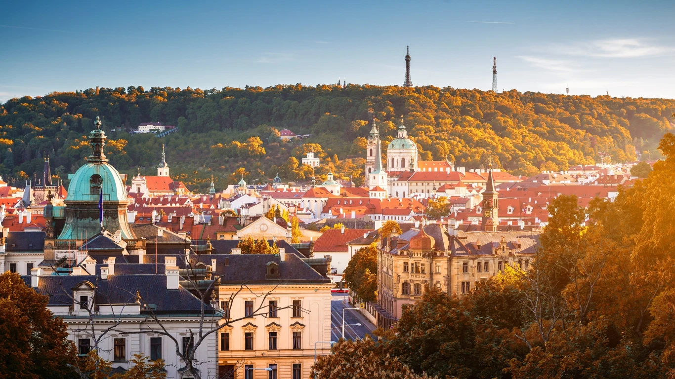 Panorama miasta z dominującymi czerwonymi dachami, barokowymi wieżami kościołów i kopułą, w tle zielone wzgórze z wieżą widokową i antenami, całość otoczona złotymi, jesiennymi drzewami.
