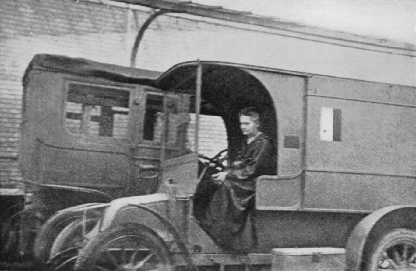 Maria Skłodowska-Curie w jej samochodzie do prześwietleń Kobieta siedząca za kierownicą zabudowanego samochodu z początku XX wieku, ubranego w długą suknię, na tle ceglanego muru.