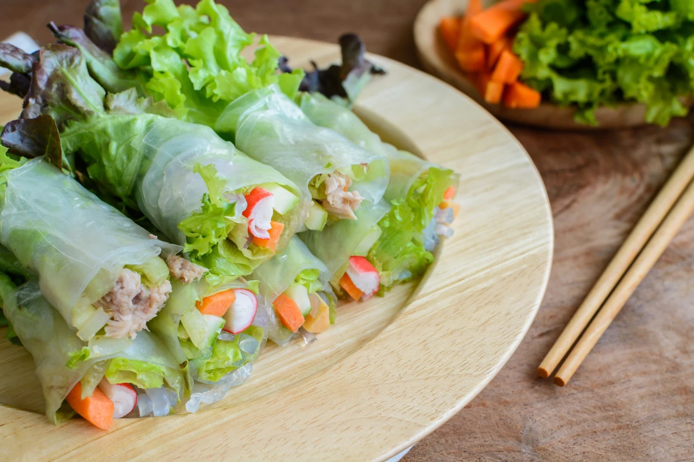 Pyszne i lekkie sajgonki bez smażenia. Idealne na kolację lub przekąskę w większym gronie Sajgonki bez smażenia z sałatą, surimi, marchewką i tuńczykiem w papierze ryżowym, na drewnianym talerzu.