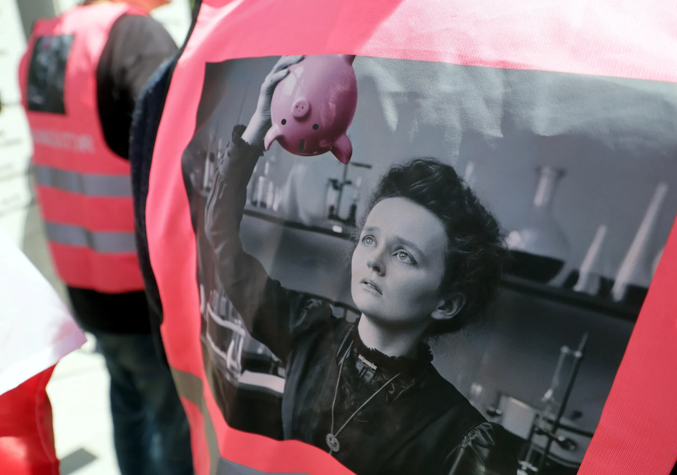 Maria Skłodowska-Curie na kurtkach protestujących pod Sejmem Różowa kamizelka z wielkim nadrukiem czarno-białego zdjęcia kobiety w laboratorium, która wznosi w górę różową świnkę-skarbonkę, podczas gdy inni uczestnicy ubrani są w podobne kamizelki.