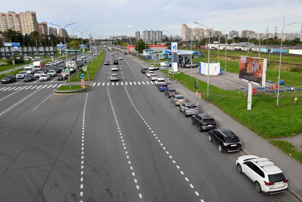 Długa kolejka samochodów oczekujących na wjazd na stację benzynową przy szerokiej, miejskiej arterii, wokół zabudowa wielorodzinna i widoczne billboardy reklamowe.
