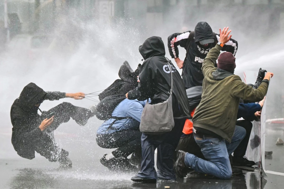 Protesty na ulicach Brukseli Grupa ludzi w ciemnych ubraniach i kapturach osłania się przed silnym strumieniem wody z armatki wodnej, próbując utrzymać równowagę i chronić się nawzajem.