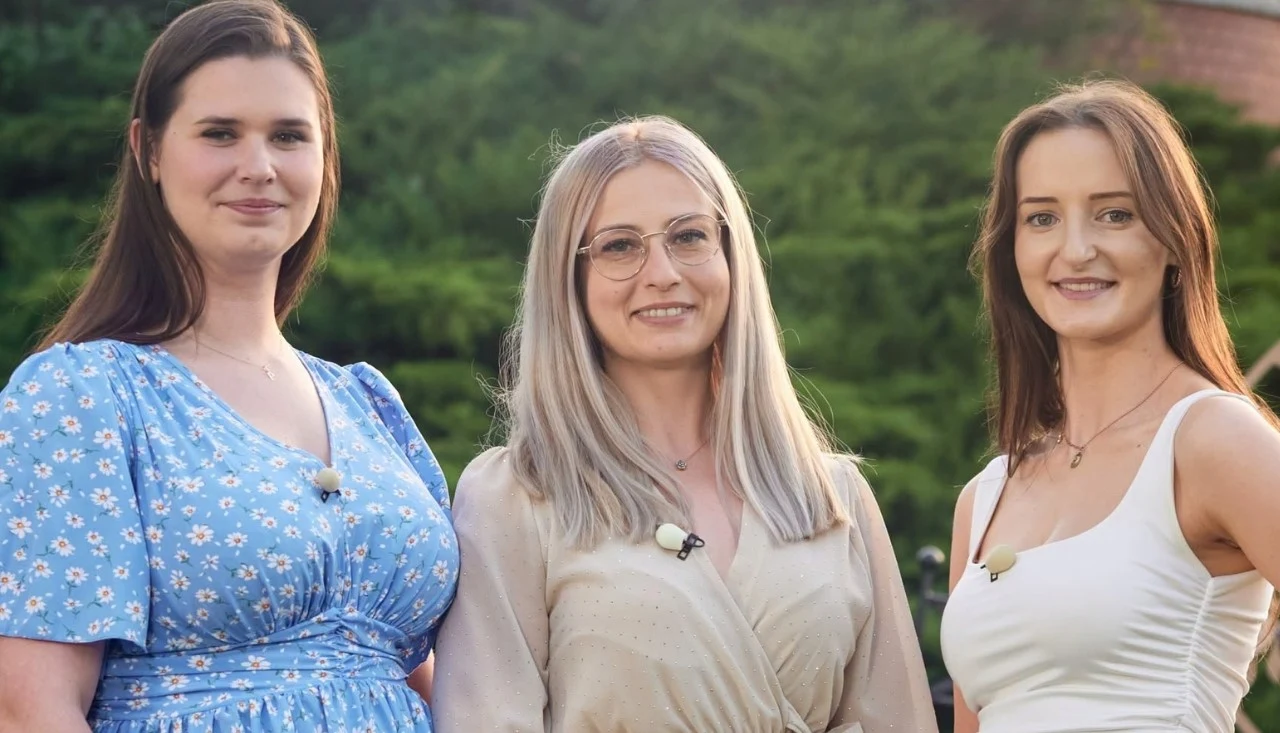 Trzy uśmiechnięte kobiety stoją obok siebie na tle zielonej roślinności, każda ubrana w letnią sukienkę i wyposażona w mikrofon przypinany do ubrania.