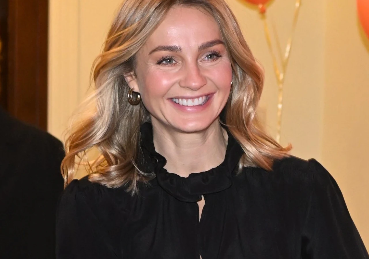 Małgorzata Socha Uśmiechnięta kobieta z blond włosami, ubrana w elegancką czarną bluzkę, stoi na tle jasnej ściany ozdobionej balonami.