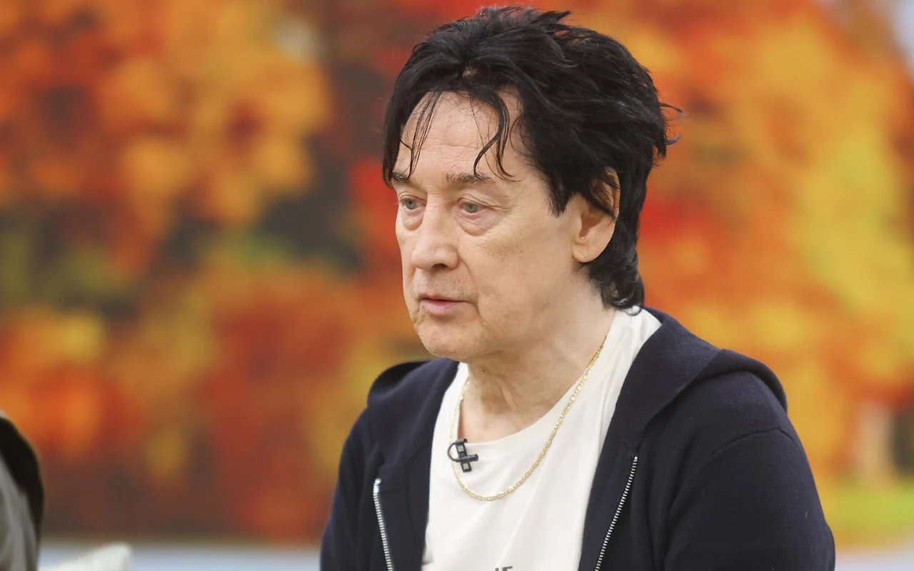 Jan Borysewicz w "halo tu polsat" Mężczyzna o ciemnych, lekko potarganych włosach, ubrany w jasny t-shirt i ciemną rozpinaną bluzę, z mikrofonem przypiętym do koszulki, na tle rozmytych jesiennych barw.