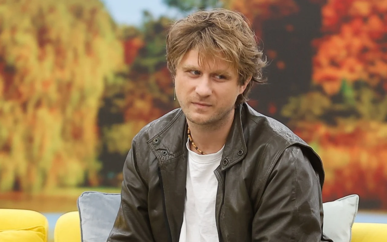 Patryk Pietrzak w studiu "halo tu polsat" Mężczyzna z jasnobrązowymi włosami w skórzanej kurtce i białej koszulce siedzi na żółtej sofie, w tle rozmyte jesienne drzewa.