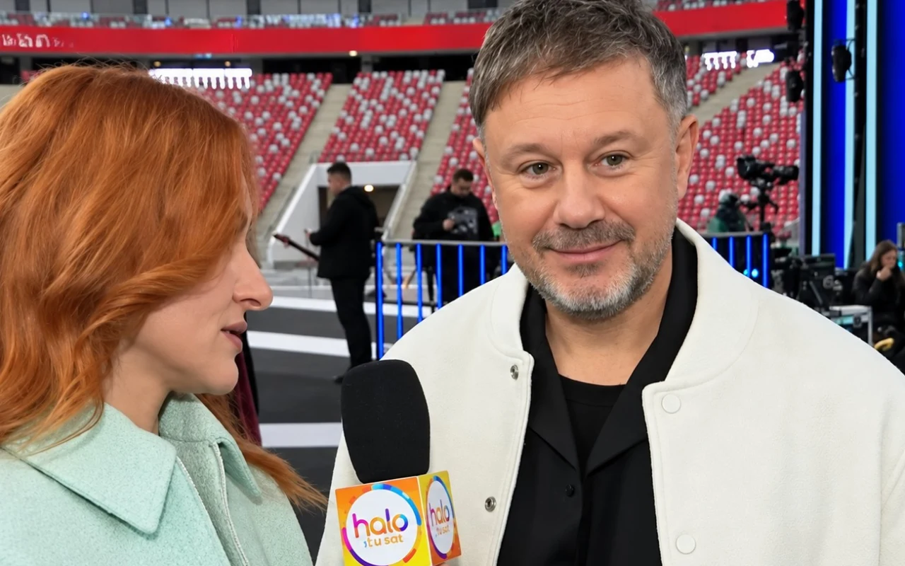 Rozmowa dwóch osób w jasnych płaszczach, z czego jedna trzyma mikrofon z logo „halo tu studio”; w tle stadion z czerwonymi krzesełkami i ekipą techniczną.
