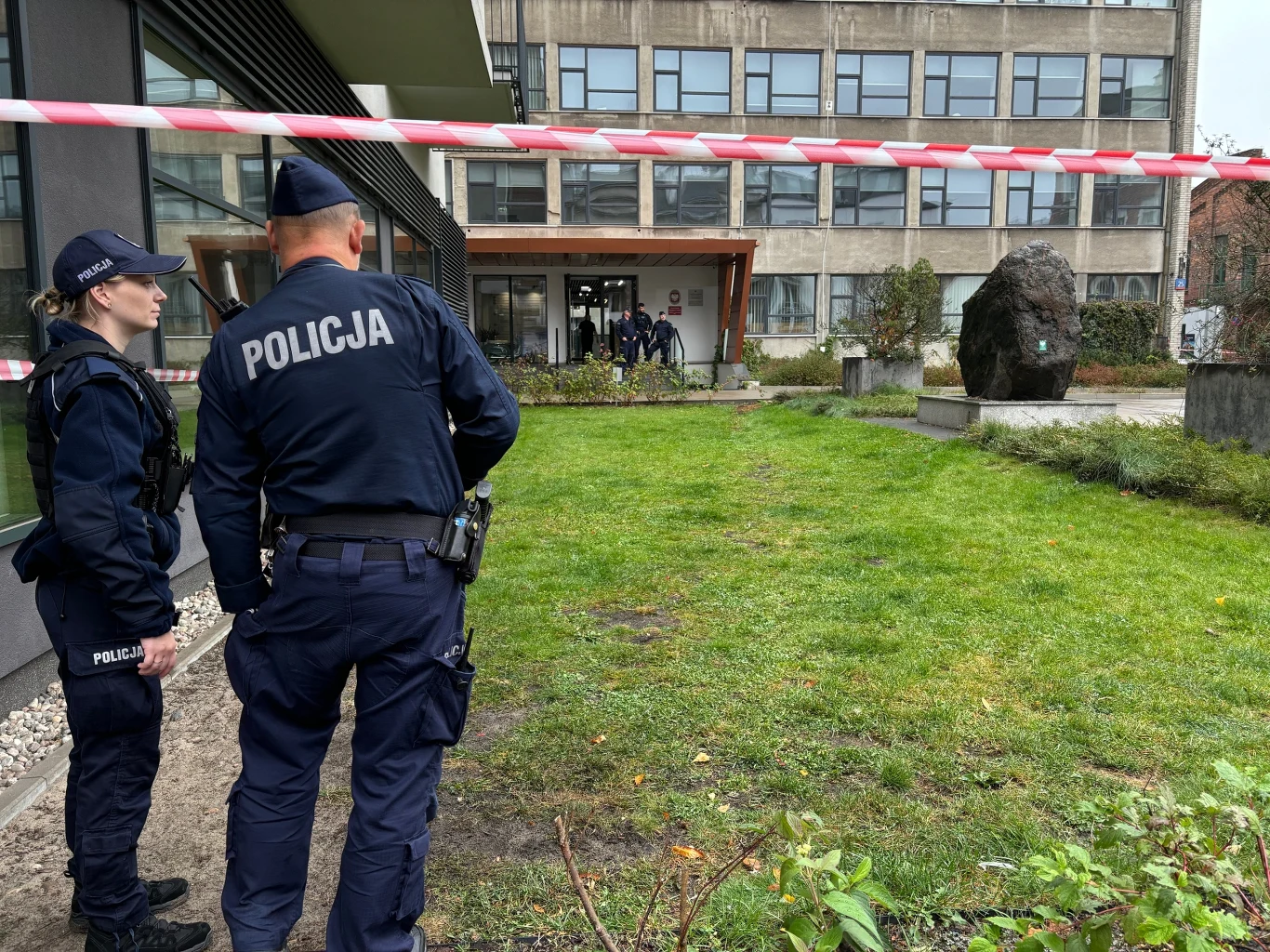 Dwóch policjantów w mundurach stoi za taśmą odgradzającą teren przed wejściem do budynku użyteczności publicznej, w tle widoczna duża bryła kamienia oraz grupa osób przy wejściu.