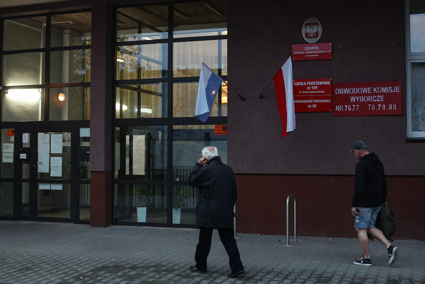 Wejście do szkoły w Krakowie z tablicami informacyjnymi, polską flagą i wyborcami wchodzącymi do środka.