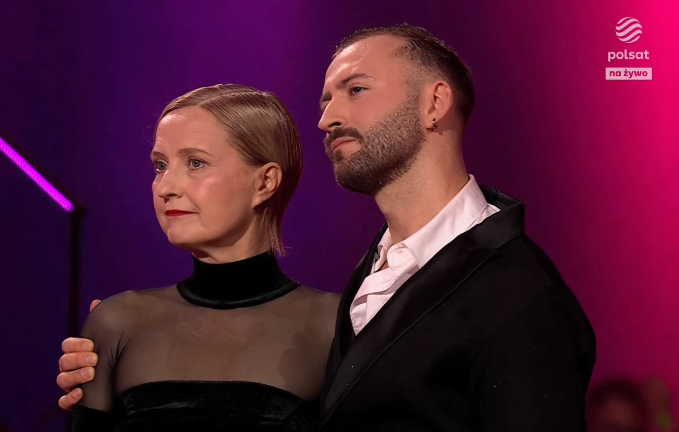 Taniec z gwiazdami - Izabela Kuna i Michał Bartkiewicz Dwie osoby stoją obok siebie, obejmując się, obie ubrane elegancko, na tle efektownego, fioletowego oświetlenia scenicznego z widocznym logo telewizji Polsat w rogu.