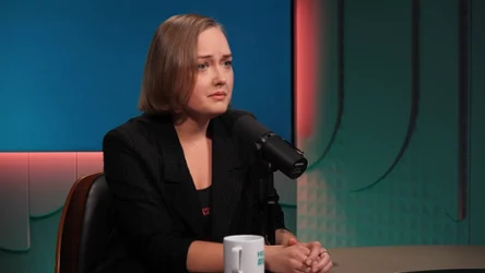 Kobieta z krótkimi, blond włosami w czarnym żakiecie siedzi przy stole w studio radiowym lub podcastowym, trzyma dłonie na blacie i mówi do mikrofonu, obok stoi biały kubek.