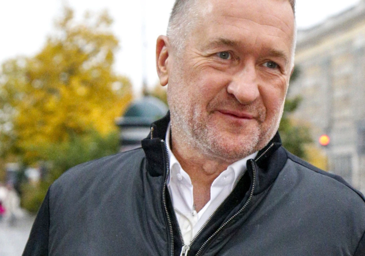 Rafał Sonik Rafał Sonik w czarnej kurtce i białej koszuli, pozujący na tle jesiennej zieleni oraz miejskiej zabudowy.