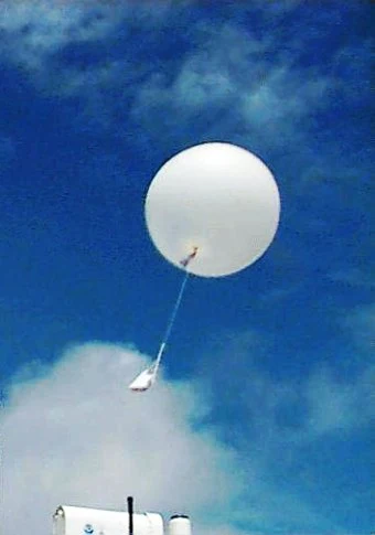 Biały balon meteorologiczny unoszący się na tle błękitnego nieba z podwieszoną aparaturą badawczą.