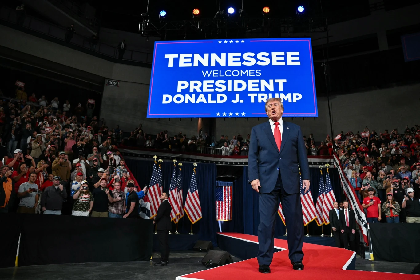 Donald Trump stoi na scenie w hali widowiskowej, za nim widoczny jest duży ekran z napisem powitalnym oraz zgromadzony tłum, ozdobiony licznymi amerykańskimi flagami.