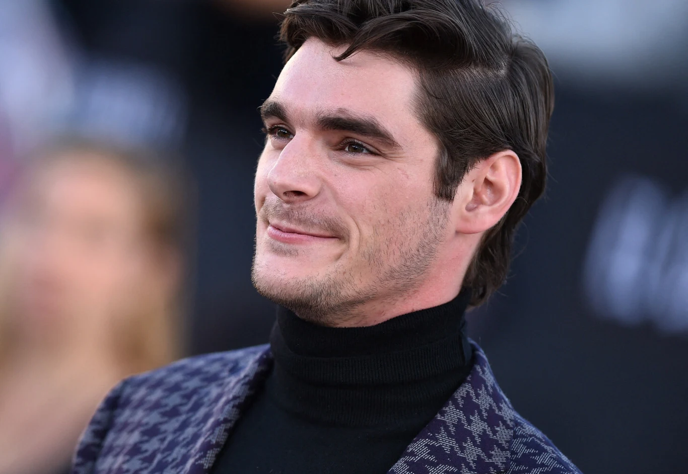 RJ Mitte Uśmiechnięty RJ Mitte, aktor znany z serialu Breaking Bad, ubrany w ciemny golf i marynarkę w kratę.