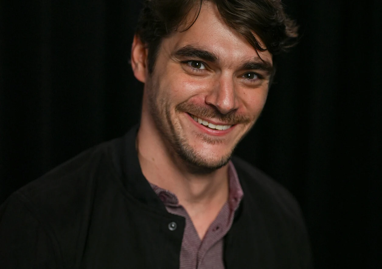RJ Mitte Uśmiechnięty RJ Mitte z ciemnymi włosami i zarostem w rozpiętej koszuli oraz ciemnej marynarce na tle czarnego tła.