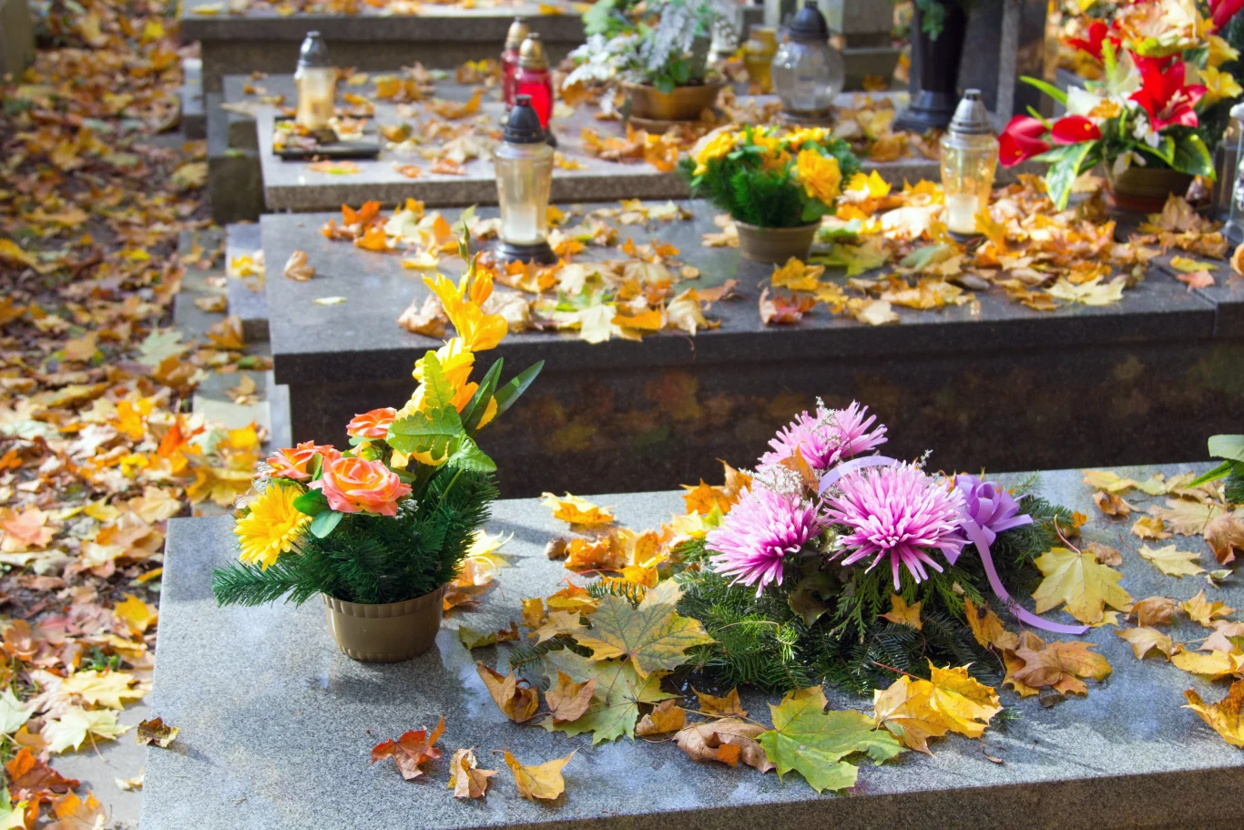 Jak zabezpieczyć kwiaty na cmentarzu przed kradzieżą? Nagrobki z kolorowymi wieńcami, kwiatami i zapalonymi zniczami na tle opadłych liści jesiennych.