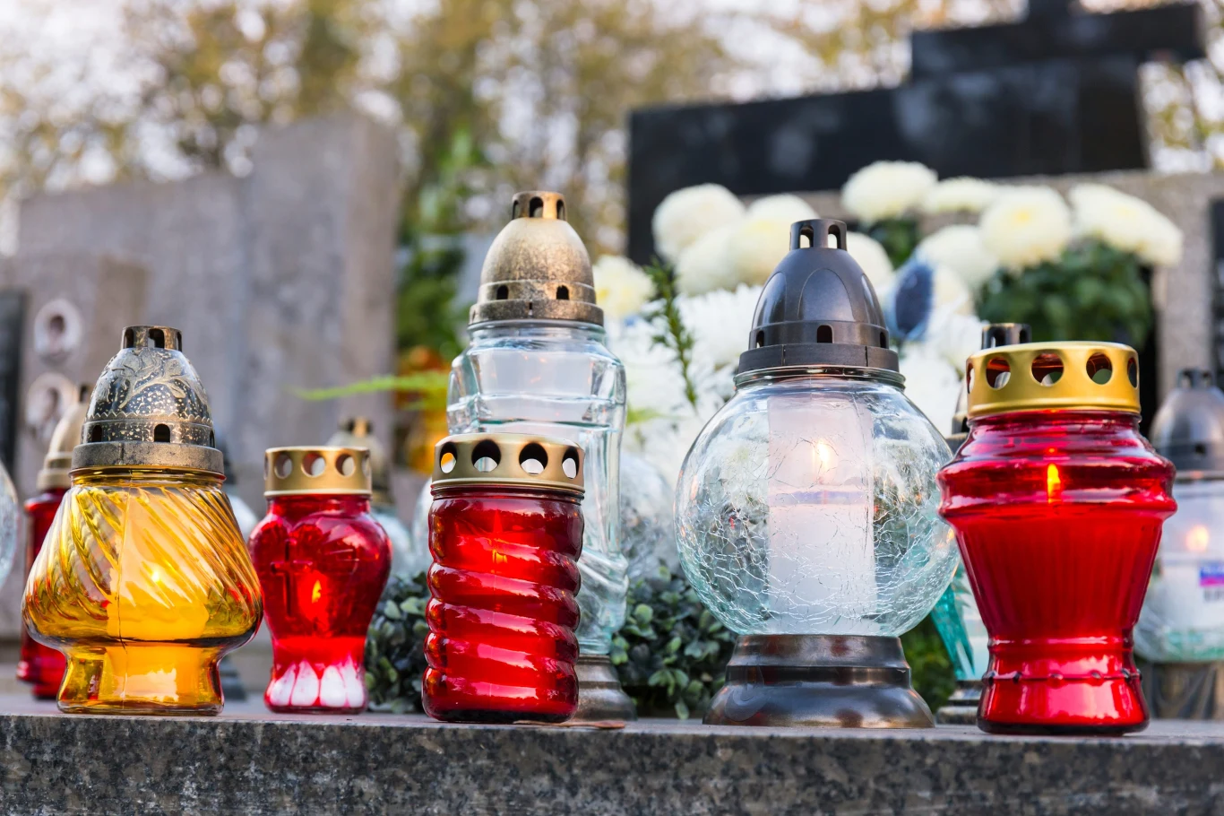 Dlaczego na grobach od wieków stawia się znicze? Kolorowe znicze ustawione na nagrobku, w tle biały bukiet kwiatów na cmentarzu.