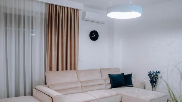 Jasny, nowoczesny salon z dużym narożnikiem w odcieniu beżowym, ciemnymi poduszkami, minimalistycznym zegarem na ścianie oraz zasłonami w ciepłych kolorach.