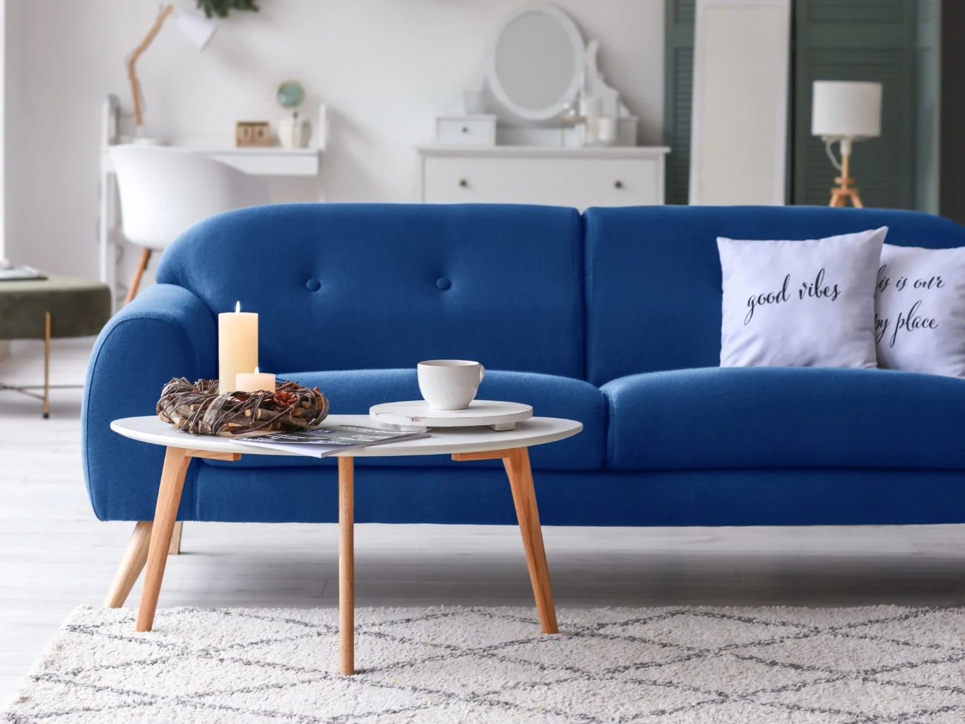 Niebieska sofa z białą poduszką, przed nią biały stolik z kawą, świecą i ozdobą z gałązek; wnętrze nowoczesne, jasne, z dywanem o geometrycznym wzorze.