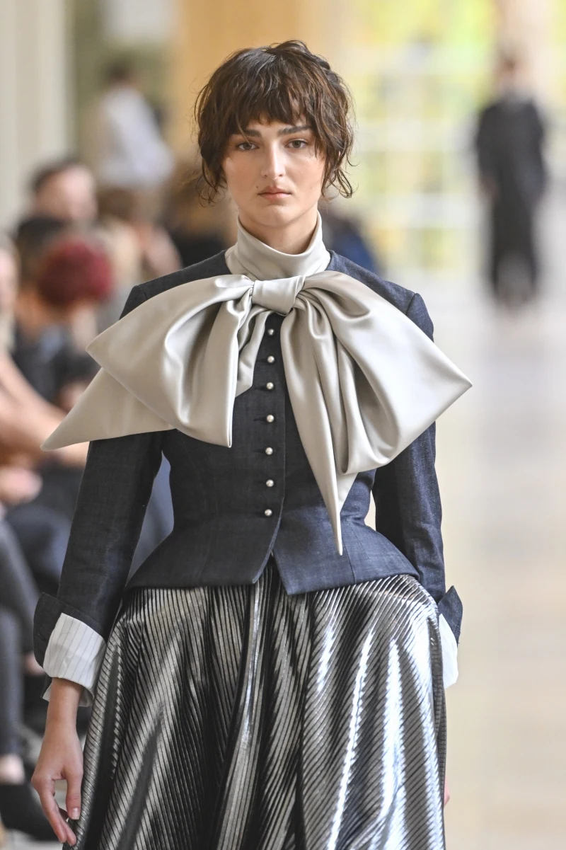 Modelka ubrana w ciemny żakiet z eleganckimi guzikami, białą koszulę z szerokimi mankietami oraz połyskującą spódnicę w prążki, z dużą, jasną kokardą ozdabiającą dekolt.