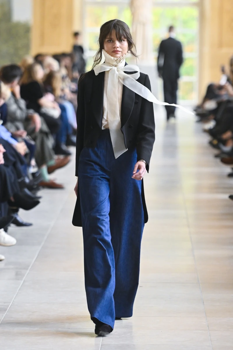 Modelka ubrana w elegancką, czarną marynarkę, białą bluzkę z szeroką kokardą przy szyi oraz szerokie, ciemnoniebieskie spodnie, idzie wzdłuż wybiegu wśród publiczności podczas pokazu mody.