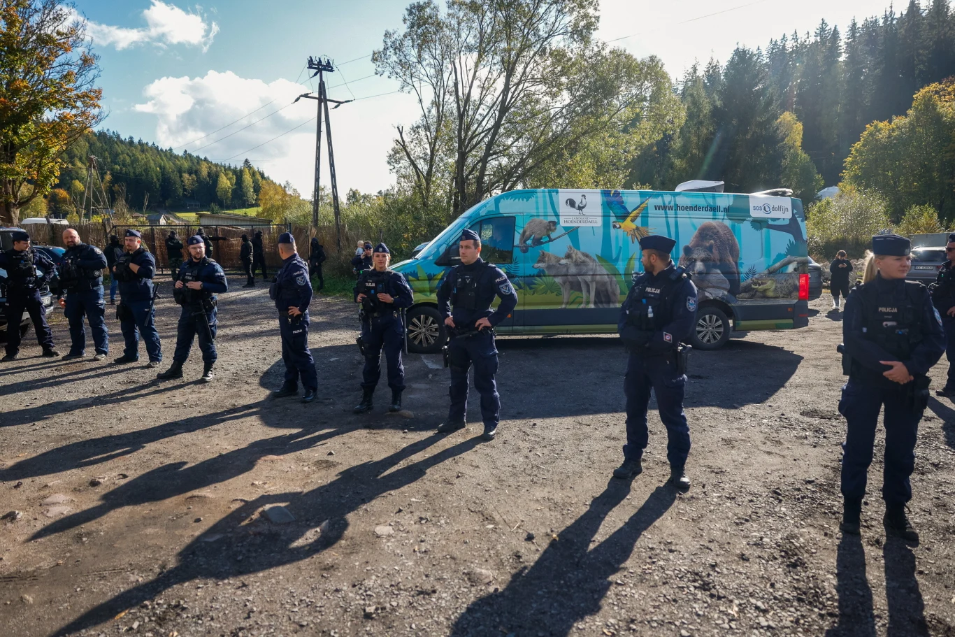 Grupa umundurowanych policjantów ustawiona w linii na żwirowym placu, w tle samochód dostawczy z kolorowymi grafikami zwierząt, otoczony przez drzewa i las.