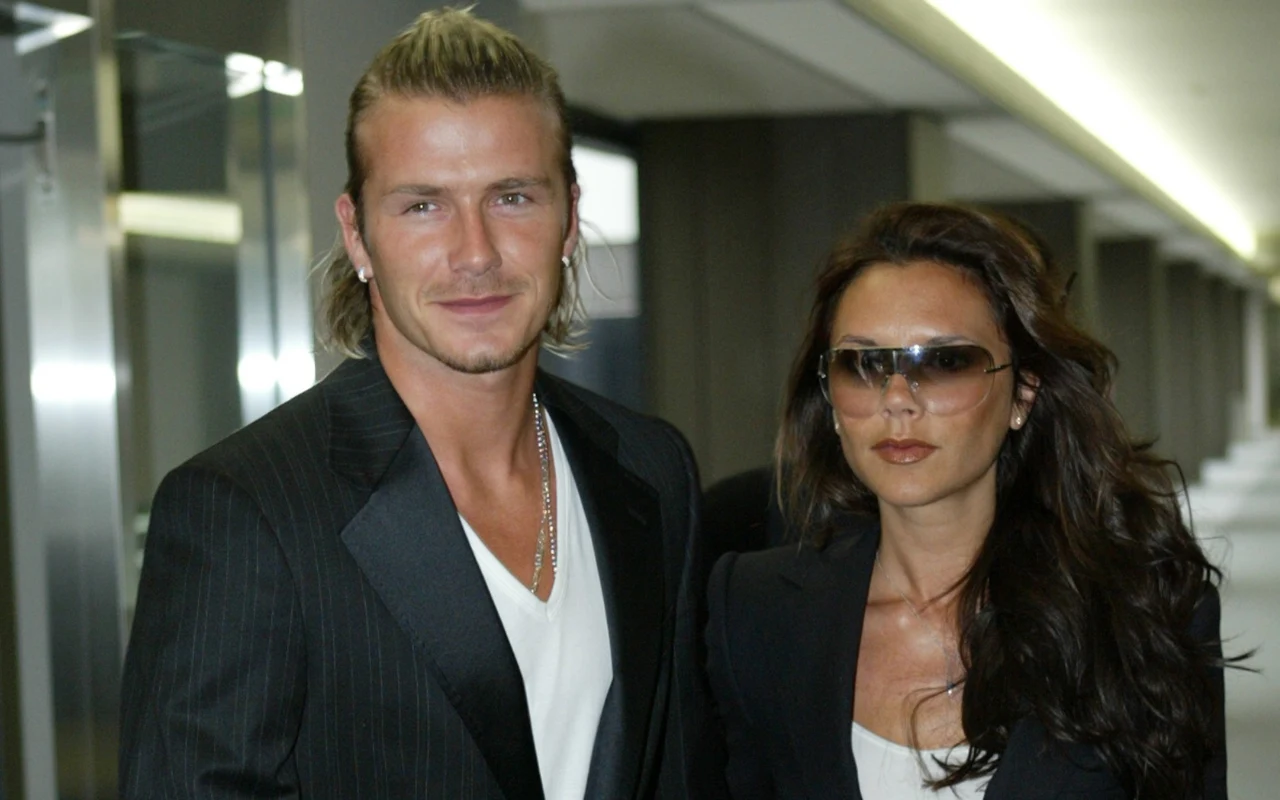 David i Victoria Beckhamowie Dwie elegancko ubrane osoby w ciemnych marynarkach, jedna z nich w okularach przeciwsłonecznych, na tle nowoczesnego, jasnego korytarza.