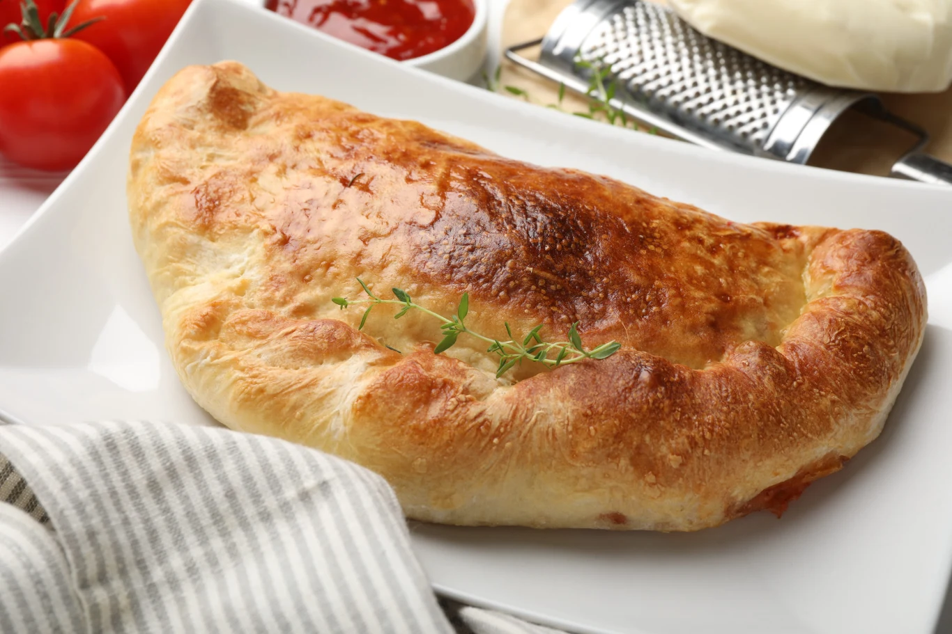 Calzone z pieczarkami, papryką i mozarellą to rodzaj pizzy w formie pieroga Calzone z pieczarkami, papryką i mozzarellą na białym talerzu z gałązką tymianku, obok miseczka sosu pomidorowego.