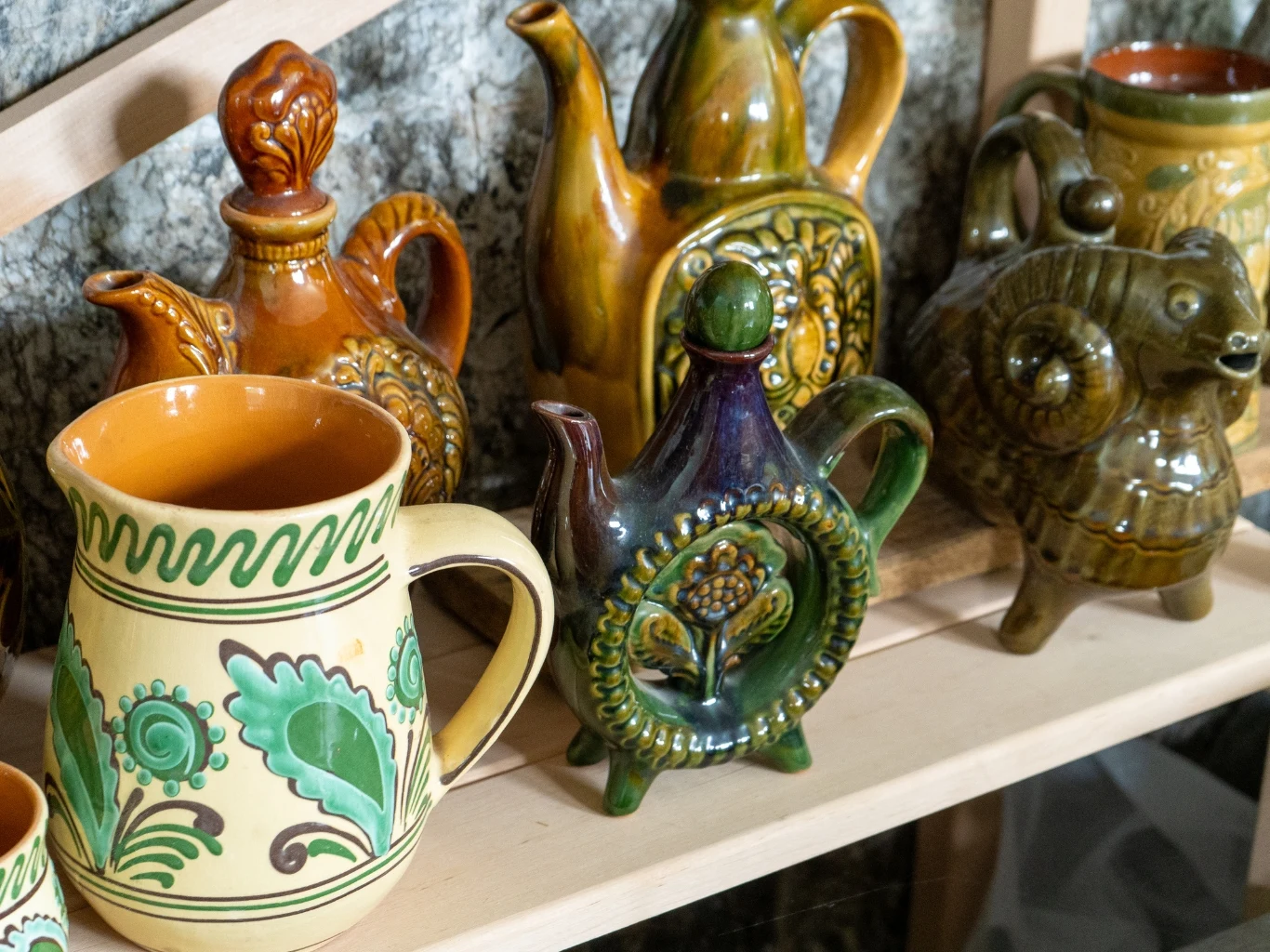 Ręcznie malowane ceramiczne dzbanki i filiżanki o bogatych barwach, zdobione tradycyjnymi wzorami, ustawione na drewnianej półce.