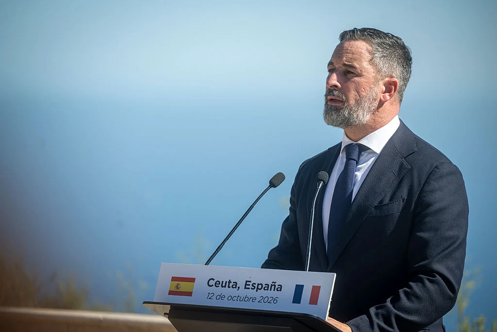 Lider hiszpańskiej Partii Vox Santiago Abascal Mężczyzna w garniturze przemawia do dwóch mikrofonów na tle nieba i morza, za mównicą z napisem Ceuta, Hiszpania oraz datą 12 de octubre 2026, obok flag Hiszpanii i Francji.