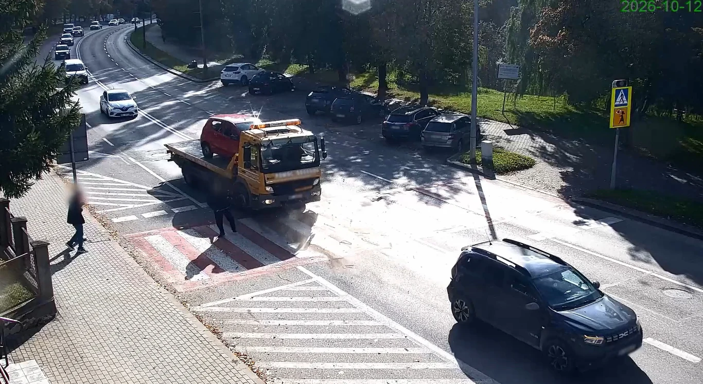 Niebezpieczne zdarzenie nagrane przez miejską kamerę. O krok od tragedii Laweta z czerwonym samochodem osobowym, piesza na przejściu, czarny SUV i inne auta na tle miejskiej ulicy.