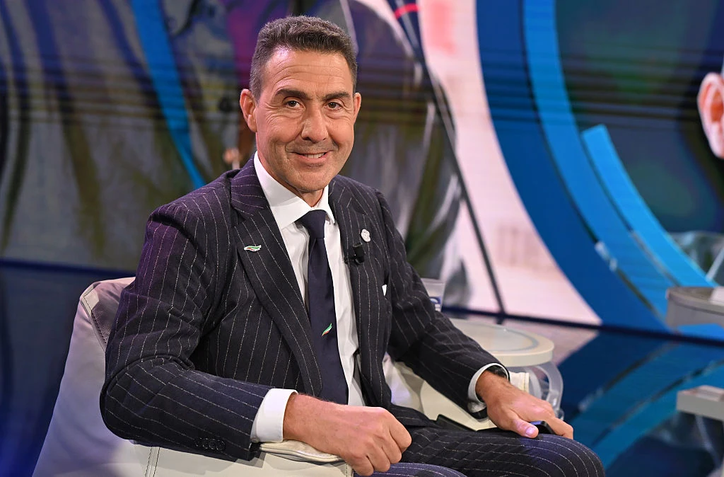 Roberto Vannacci Mężczyzna w eleganckim, ciemnym garniturze w cienkie paski siedzący na jasnym fotelu, w tle nowoczesna, kolorowa scenografia telewizyjna.