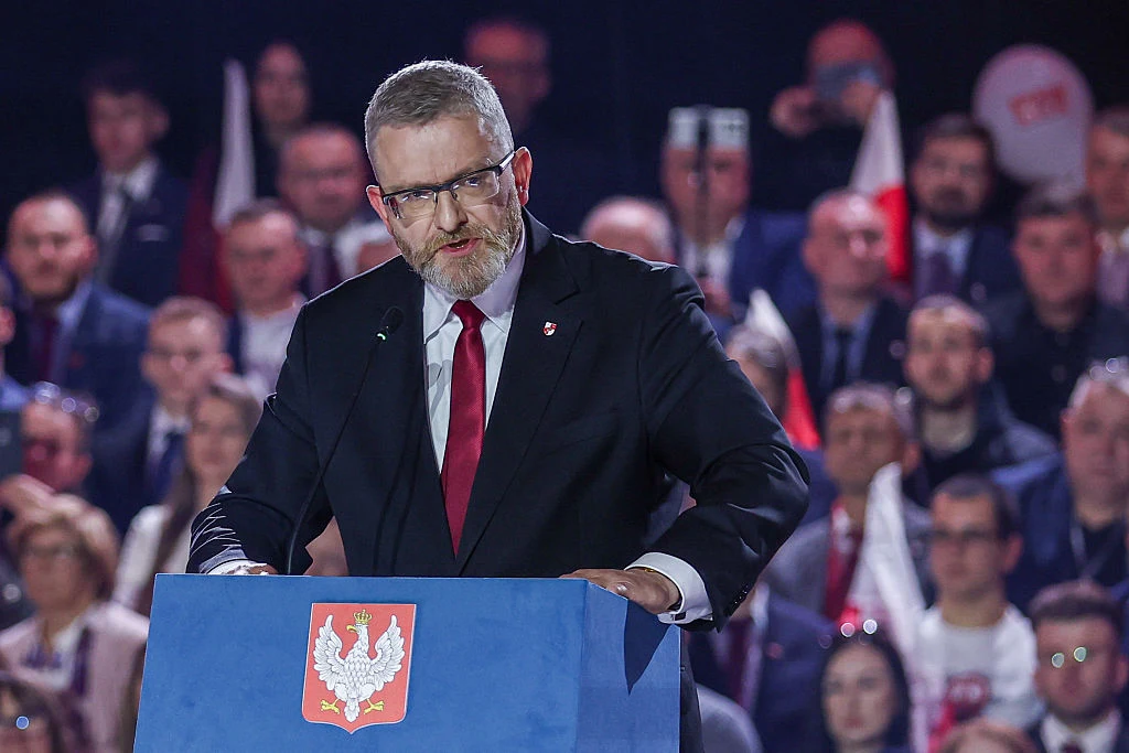Grzegorz Braun Mężczyzna w garniturze z czerwonym krawatem przemawia zza mównicy z godłem Polski, w tłumie ludzi trzymających biało-czerwone flagi.