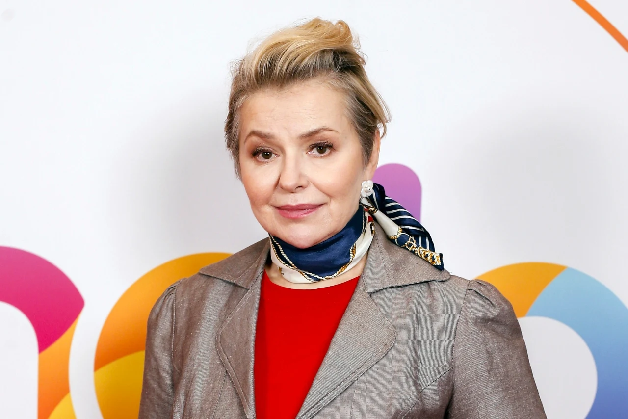 Aleksandra Konieczna jest wielbicielką apaszek, często wzbogaca swe stylizacje wzorzystymi chustkami Kobieta z blond upiętymi włosami, ubrana w szarą marynarkę, czerwoną bluzkę oraz apaszkę zawiązaną na szyi, pozuje na tle kolorowej grafiki.