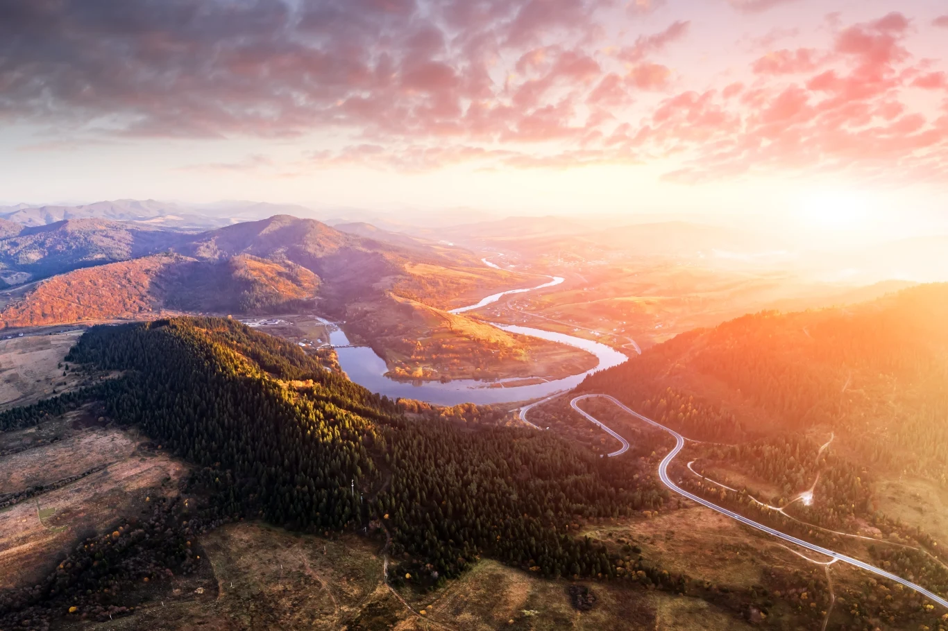 Panorama górskiego krajobrazu z wijącą się rzeką przecinającą doliny, otoczoną przez zalesione wzgórza i oświetloną promieniami zachodzącego słońca.