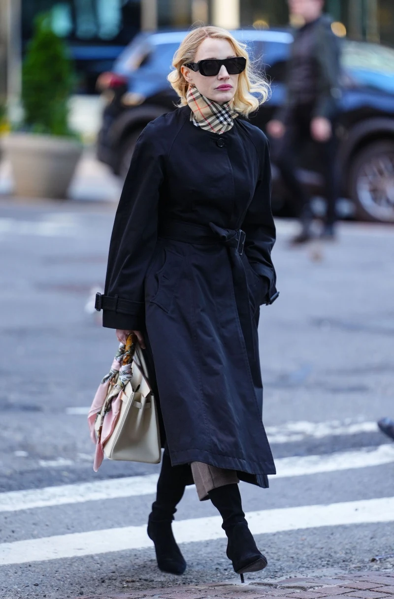 Jessica Chastain i jej torebka ozdobiona apaszką Kobieta w czarnym płaszczu, czarnych butach na obcasie i dużych okularach przeciwsłonecznych, z apaszką w kratę i jasną torbą zawiązaną kolorową chustą idzie przez przejście dla pieszych.