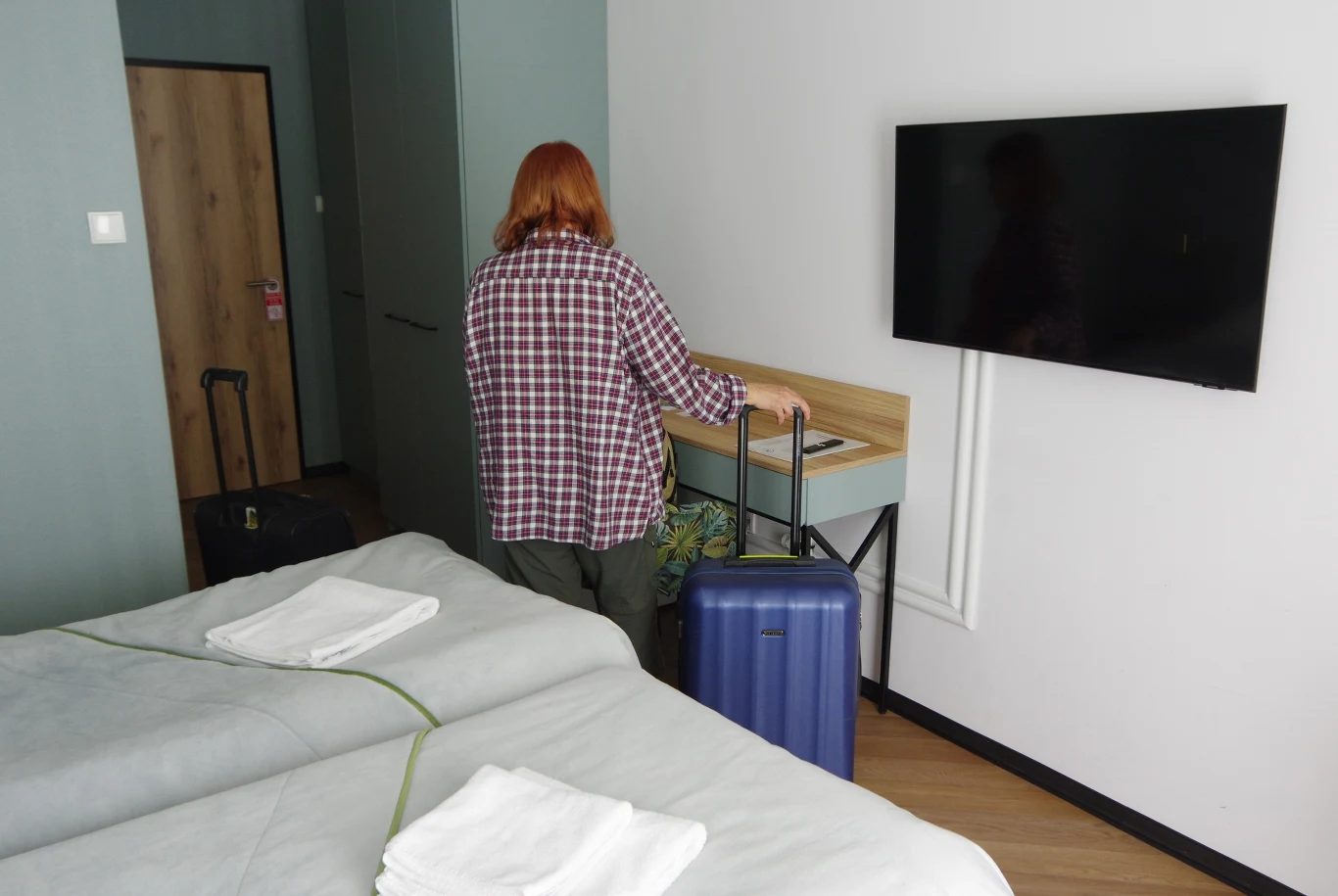 Jak przyspieszyć kolejkę do sanatorium? Kobieta z rudymi włosami ustawia niebieską walizkę przy biurku w pokoju hotelowym z dwoma łóżkami i telewizorem